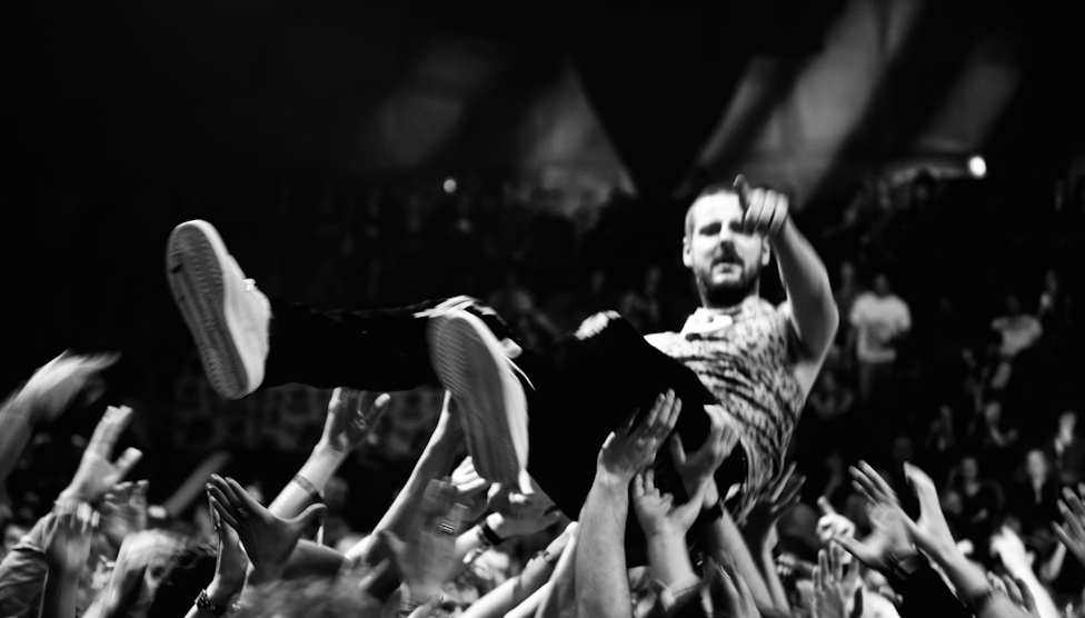 A white man wearing a t shirt, aged in his 30s, is crowdsurfing on top of many people's hands. He points at the camera.