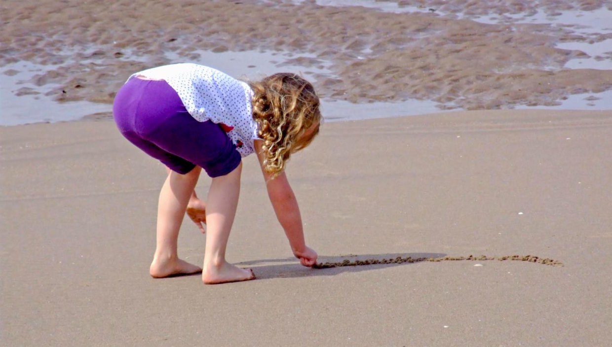 A small girl is bent over on a beach, drawing a line in the sand