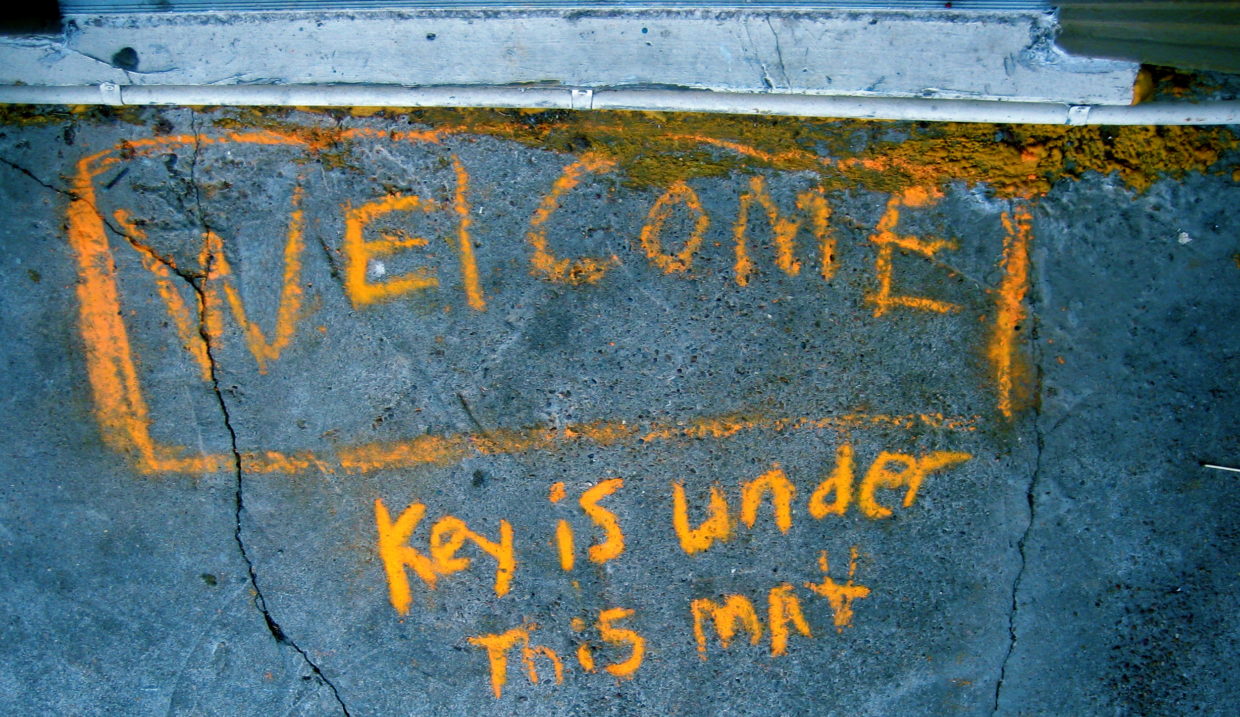 Written in yellow chalk on a sidewalk are the words "Welcome. Key is under this mat."