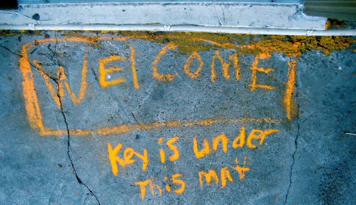 Written in yellow chalk on a sidewalk are the words "Welcome. Key is under this mat."