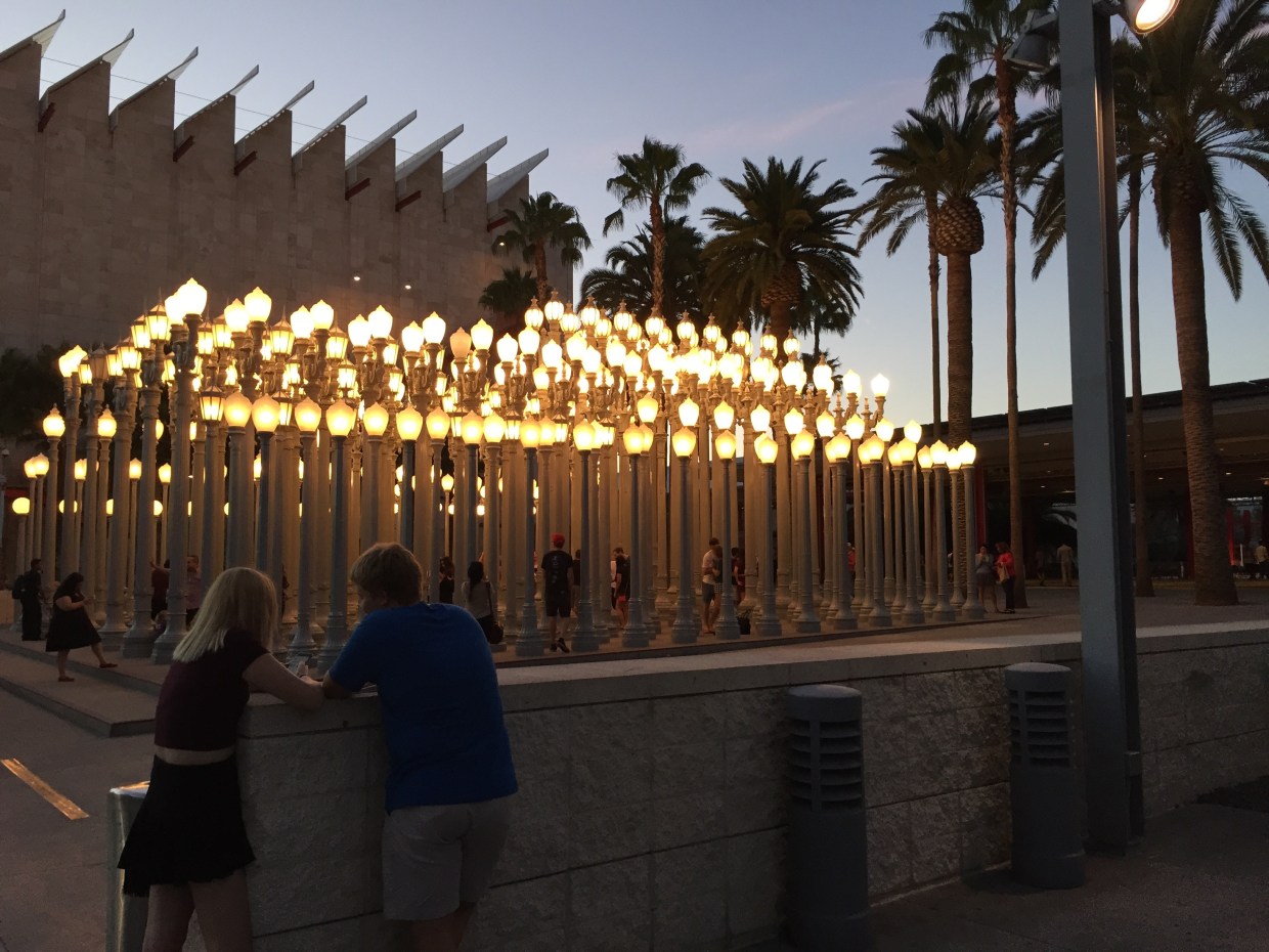 The Stanley Kubrick exhibit at Los Angeles County Museum of Art. It looks like a series of ornate, lit street lamps with people walking among them. There are palm trees, and two people in the foreground talking. It is dusk.