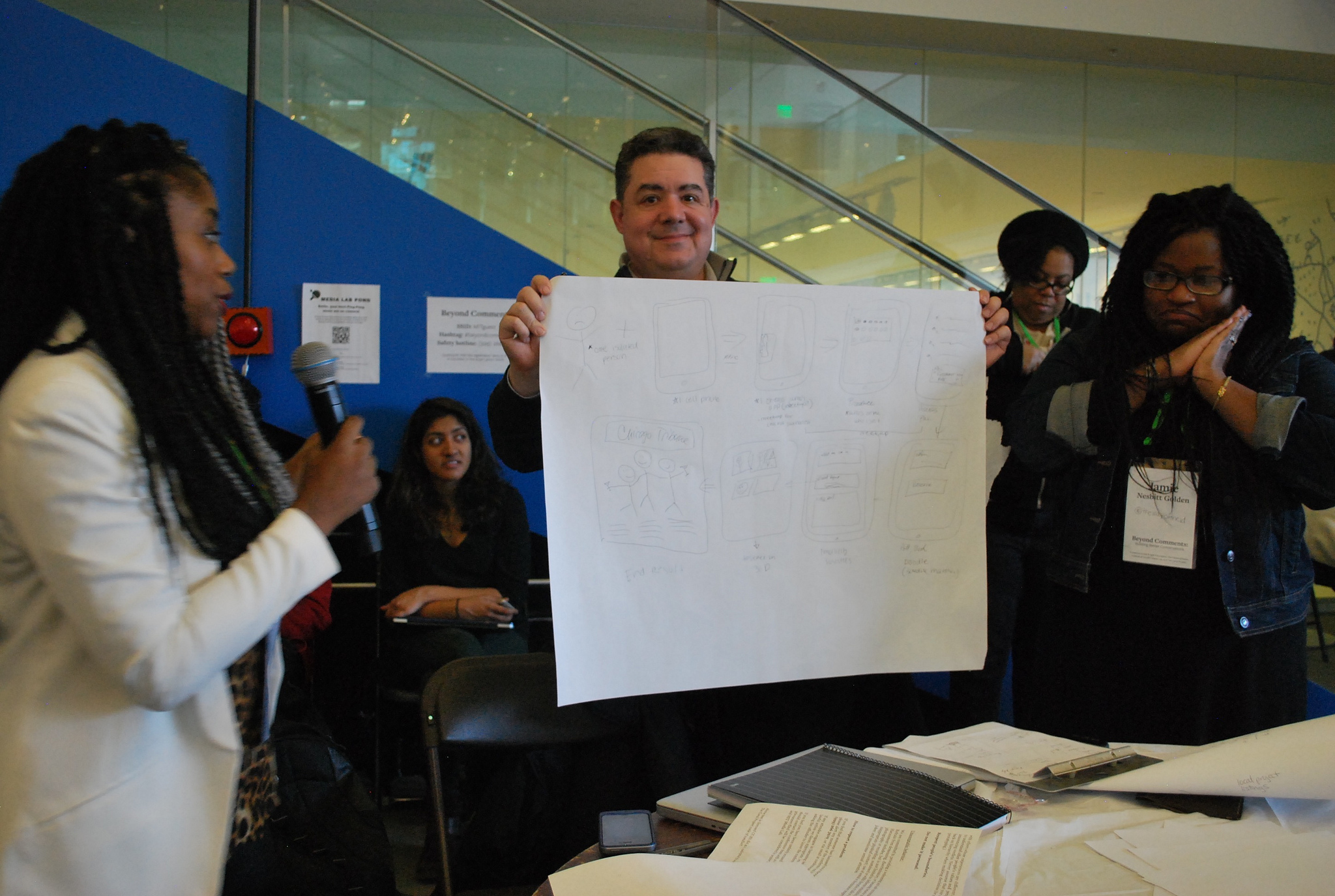 [IMAGE] A paper prototype presentation from Beyond Comments at the MIT Media Lab. Three people stand in front of a table with the person in the center holding a large piece of paper containing rough sketches. Behind them are various presenters. The person on the left has a microphone.
