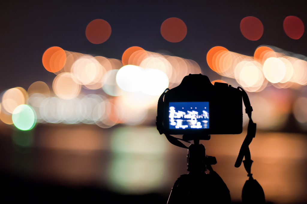 [IMAGE] Nighttime in a city, a digital camera in the foreground is on a tripod, with indistinct lights on its screen. In the background are city lights that are out of focus.