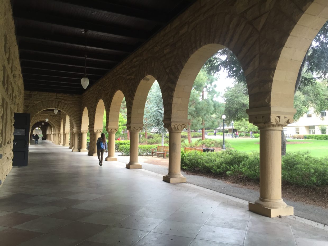 [IMAGE] A long, covered walkway with arches opening into a green courtyard. One woman is walking away from the camera in the distance.