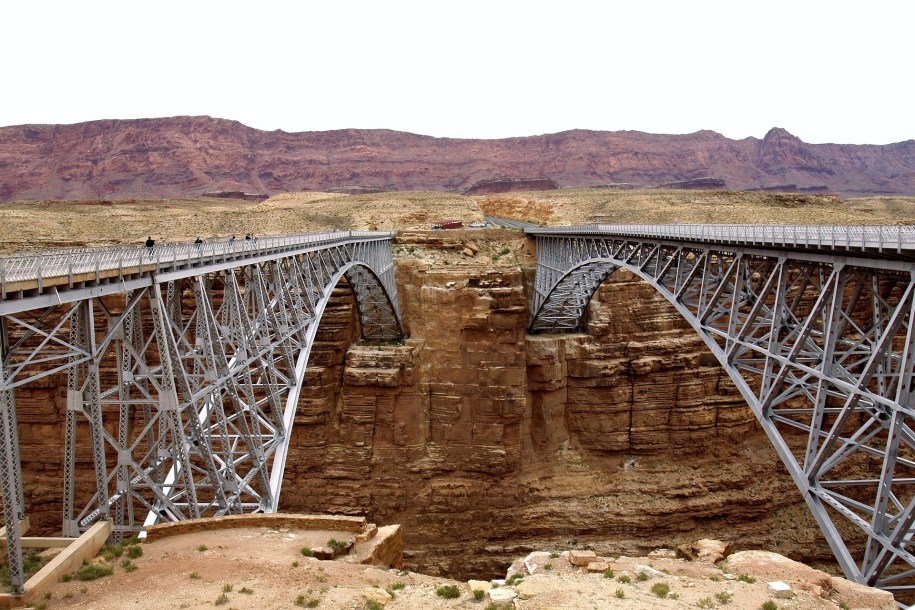 [IMAGE] A photograph taken on one side of a canyon showing two iron bridges close to each other, spanning the canyon. Small figures stand on the left bridge.