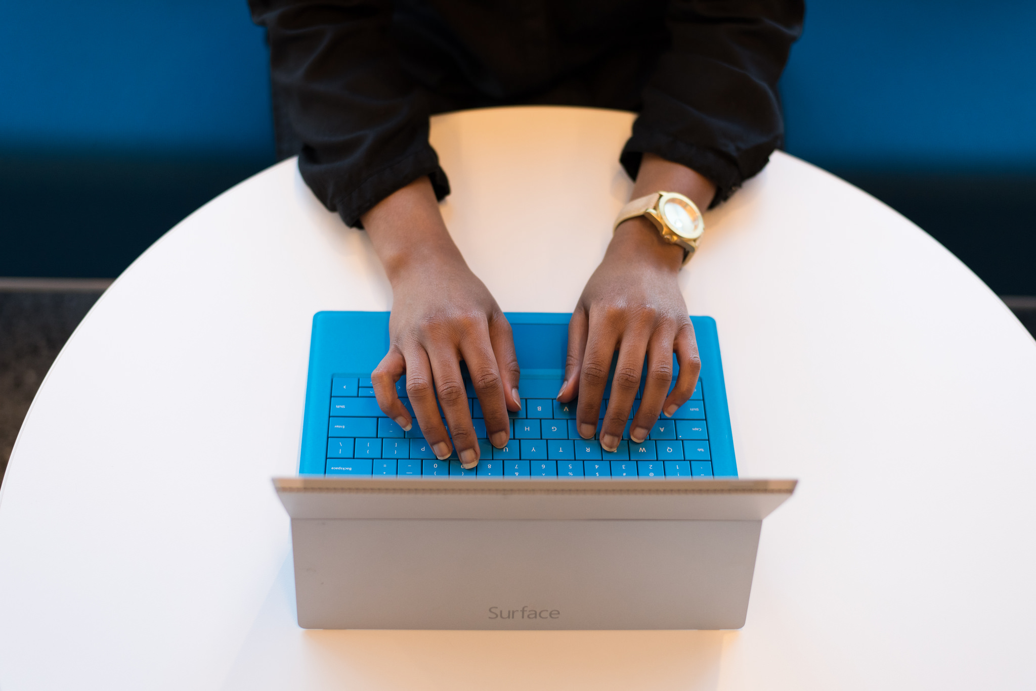 [IMAGE] A pair of dark-skinned hands wearing a gold watch are typing on a blue laptop keyboard