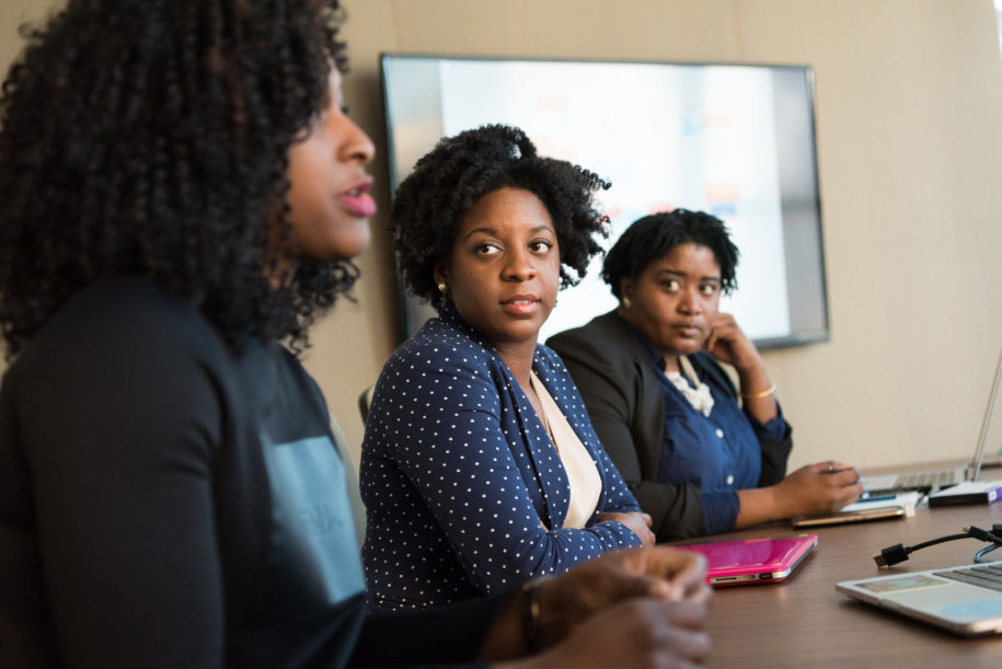 Three black women are seated at a table with computers in front of them. One is speaking, the others are listening.